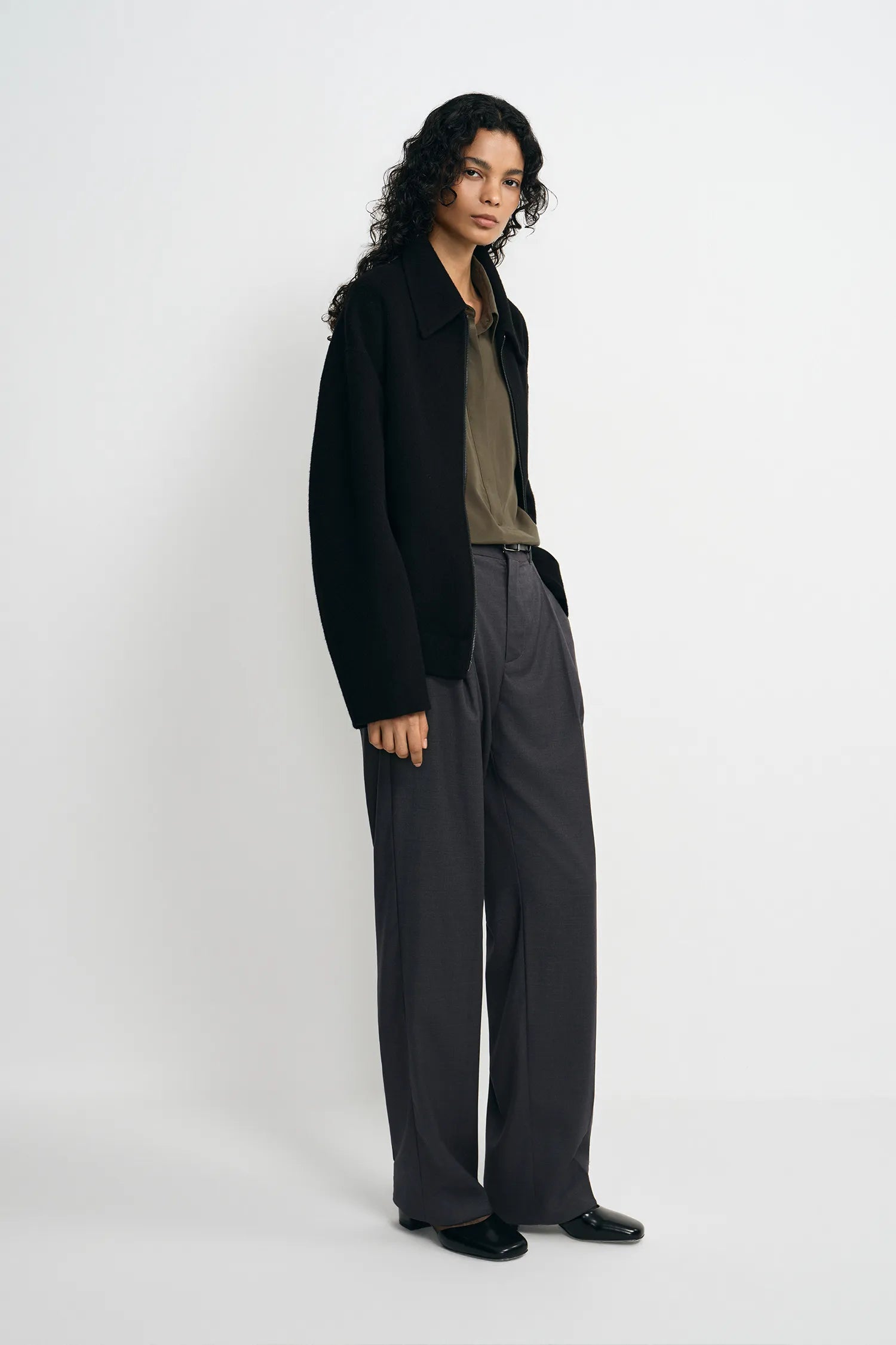A model wearing a black zip-front jacket, open, pairing an olive collared shirt and charcoal pleated trousers, side view.