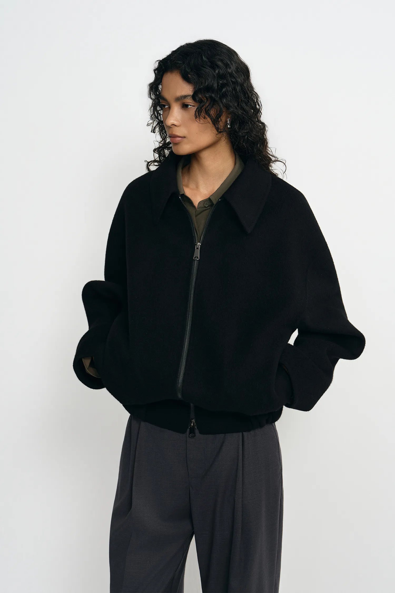 A model wearing a black zip-front jacket zipped up to the collar, pairing an olive collared shirt, turned slightly to the side.