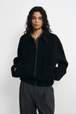 A model wearing a black zip-up jacket with a wide collar and ribbed cuffs, paired with dark gray trousers, shot against a plain light gray background.