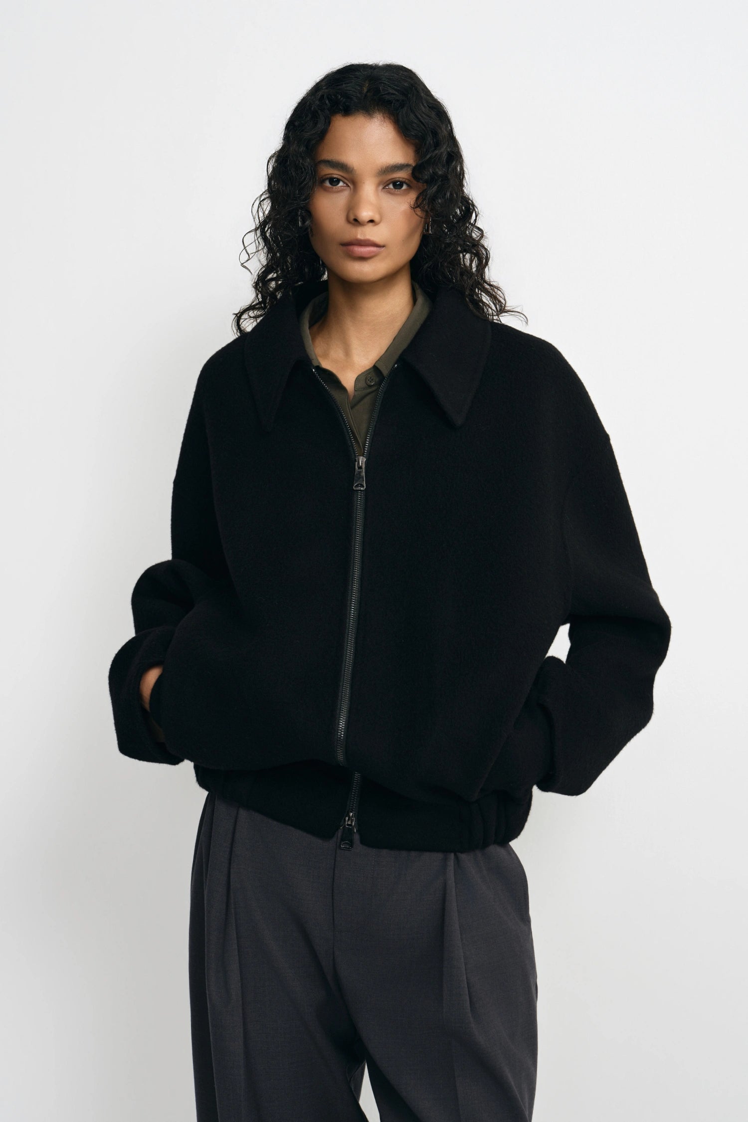 A model wearing a black zip-up jacket with a wide collar and ribbed cuffs, paired with dark gray trousers, shot against a plain light gray background.