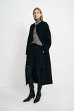 A model wearing a black collarless midi coat with a self-tie belt, pairing a gray turtleneck top, black shorts, and black knee-high boots, turned slightly to the side.