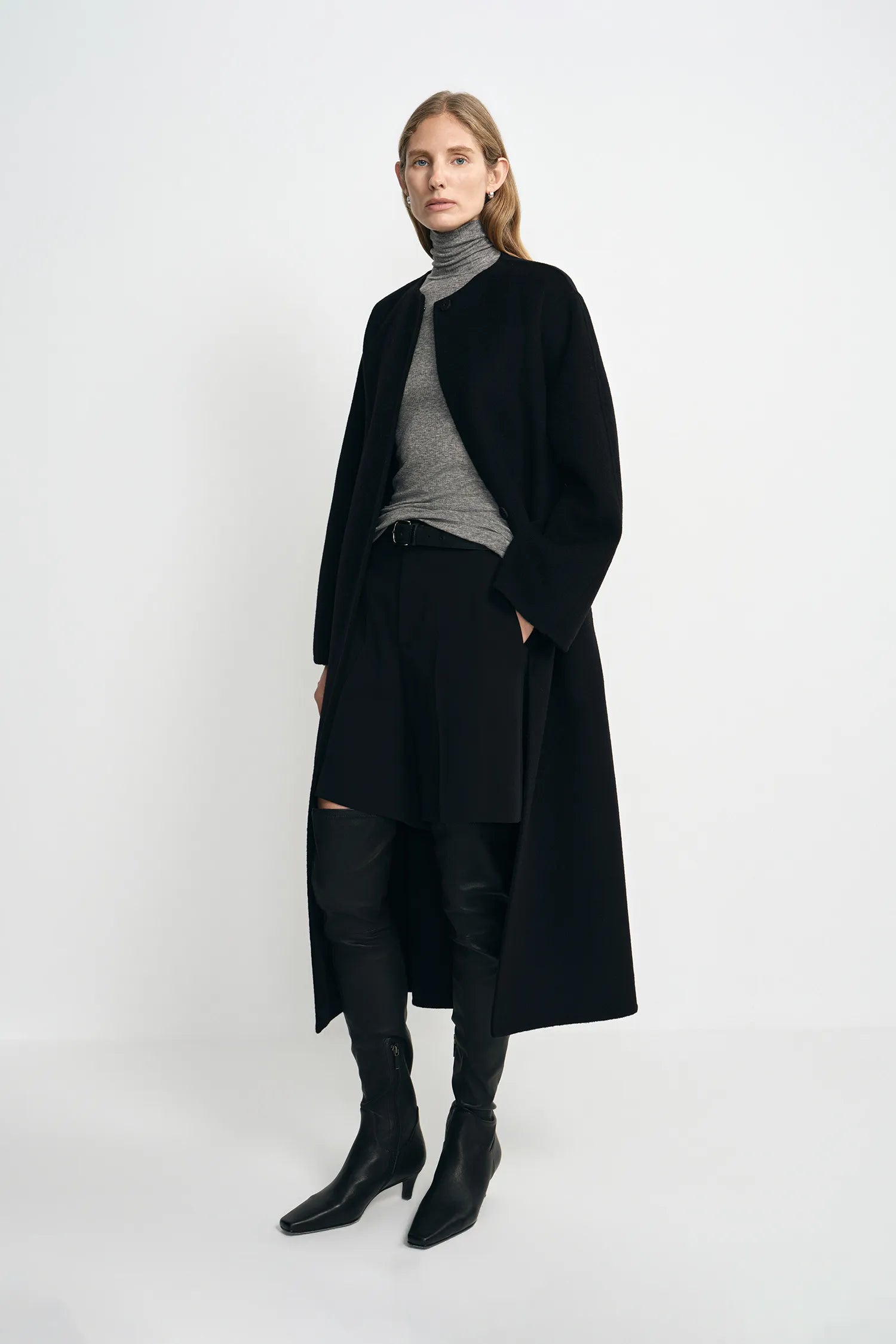 A model wearing a black collarless midi coat with a self-tie belt, pairing a gray turtleneck top, black shorts, and black knee-high boots, turned slightly to the side.