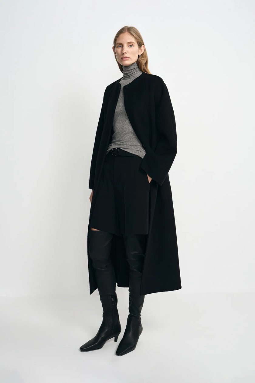 A model wearing a black collarless midi coat with a self-tie belt, pairing a gray turtleneck top, black shorts, and black knee-high boots, turned slightly to the side.