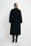 A model wearing a black collarless midi coat with a self-tie belt, pairing black knee-high boots, back view.