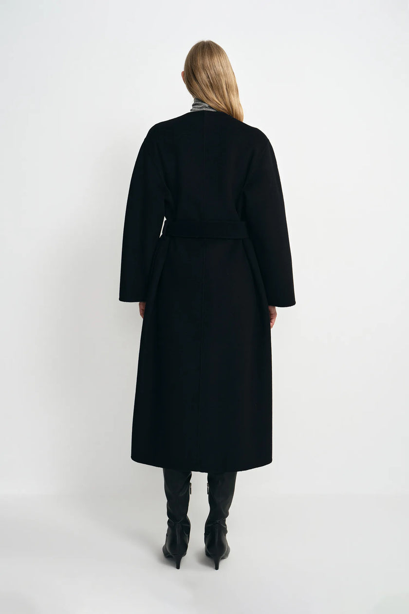 A model wearing a black collarless midi coat with a self-tie belt, pairing black knee-high boots, back view.
