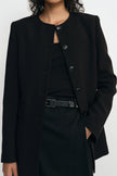 A close-up of a model wearing a black structured jacket with a round neckline, front buttons, and flap pockets, paired with a black top and skirt.