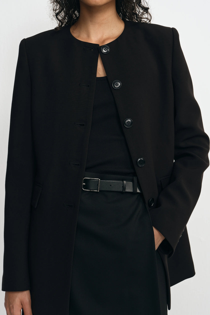 A close-up of a model wearing a black structured jacket with a round neckline, front buttons, and flap pockets, paired with a black top and skirt.