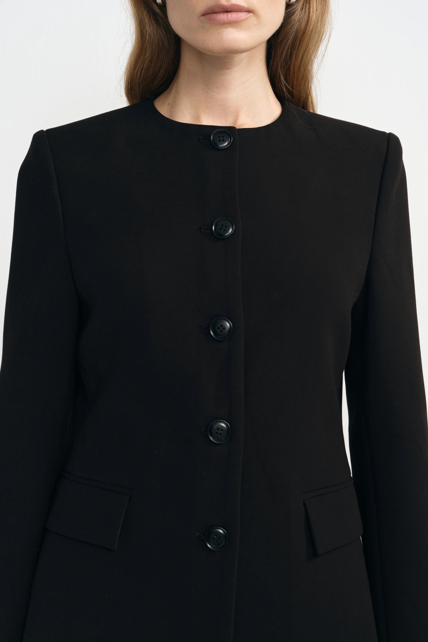 A close-up of a model wearing a black longline coat with a round neckline, a vertical row of black buttons down the front, and flap pockets near the waist.