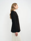 A woman shown in side profile wearing a black long-sleeve blazer over a white pleated skirt, photographed against a clean white studio background.