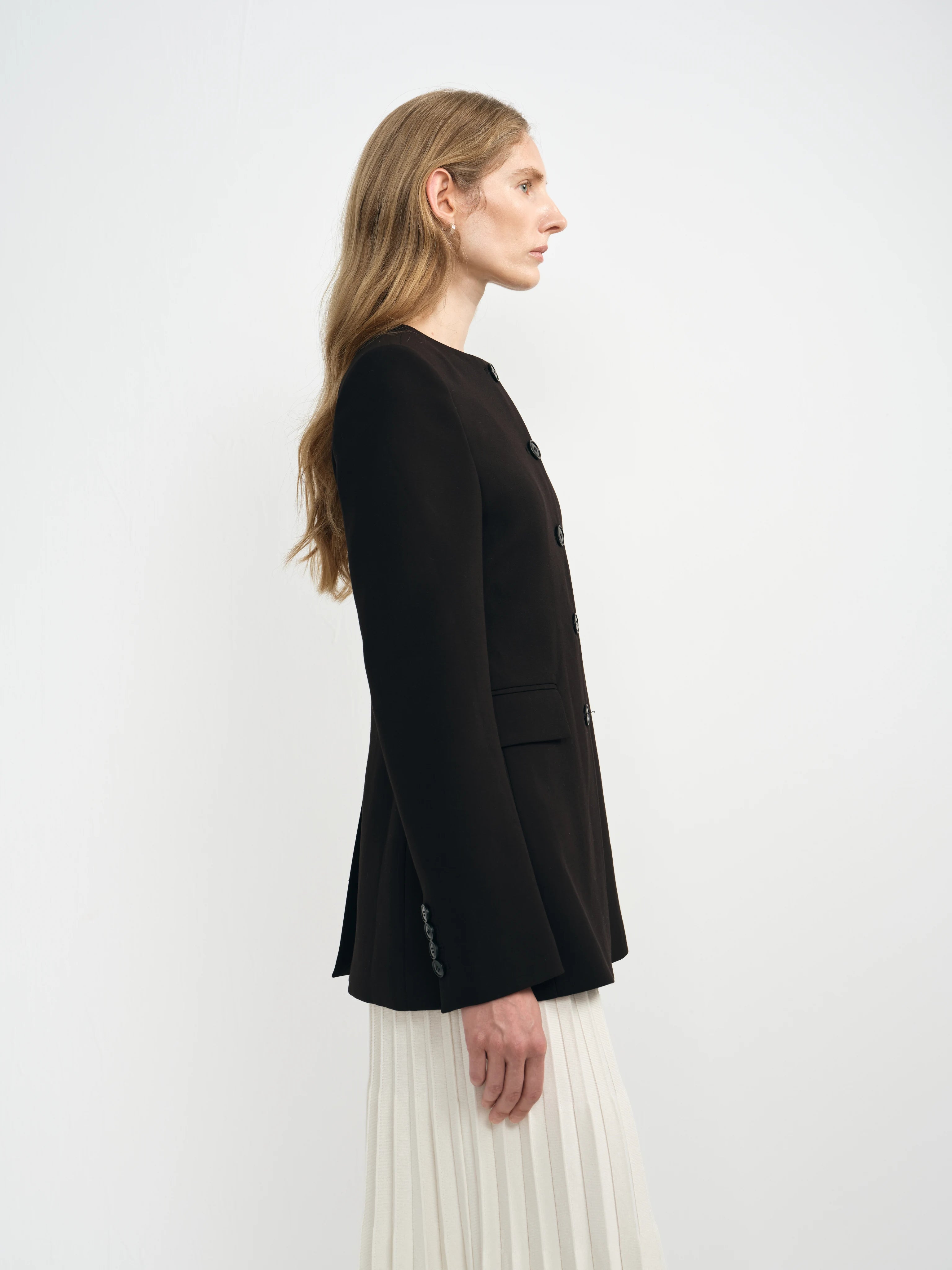 A woman shown in side profile wearing a black long-sleeve blazer over a white pleated skirt, photographed against a clean white studio background.