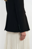 Close-up of a person wearing a black blazer with three dark buttons on the cuff and an off-white pleated skirt, against a plain light background.