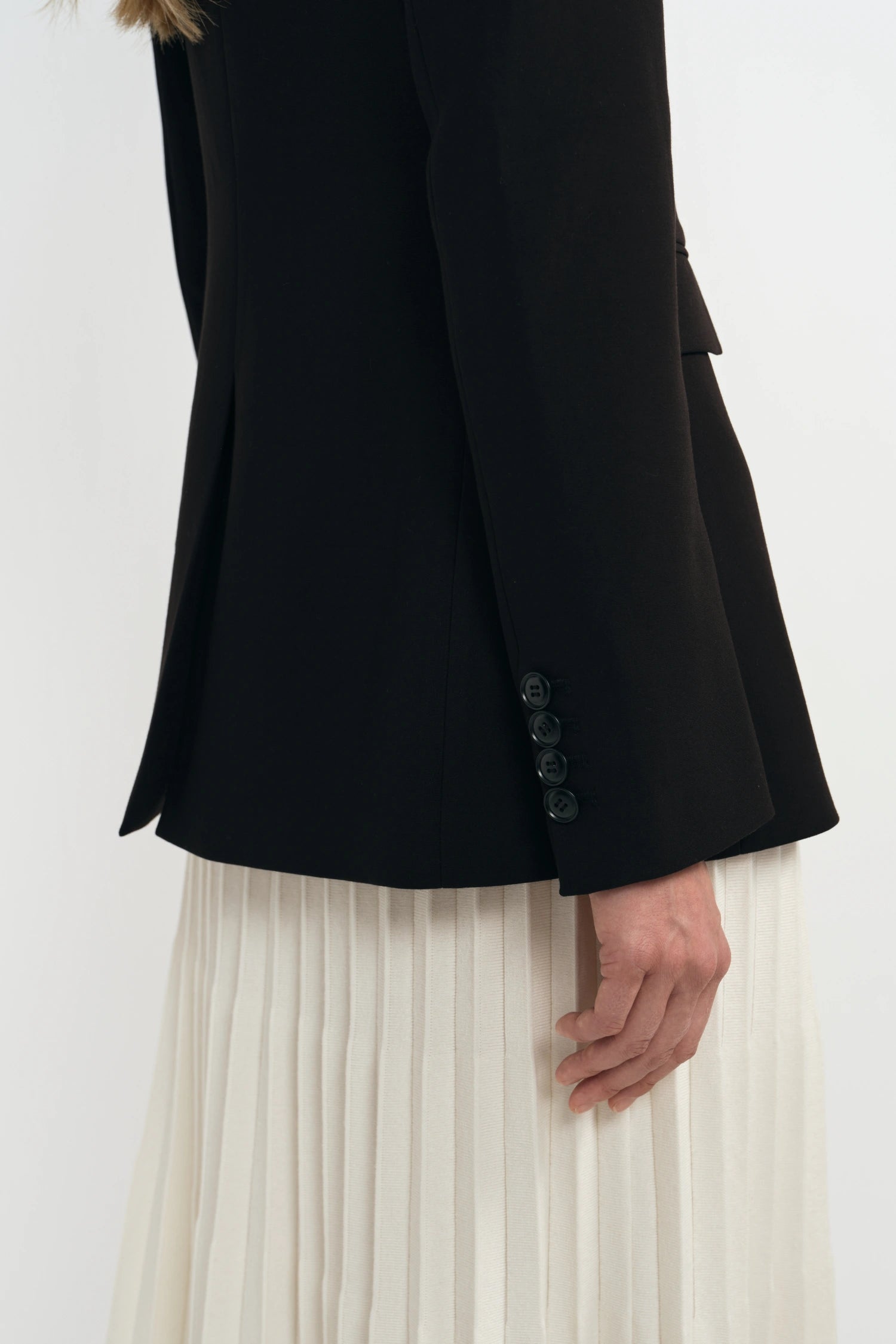 Close-up of a person wearing a black blazer with three dark buttons on the cuff and an off-white pleated skirt, against a plain light background.