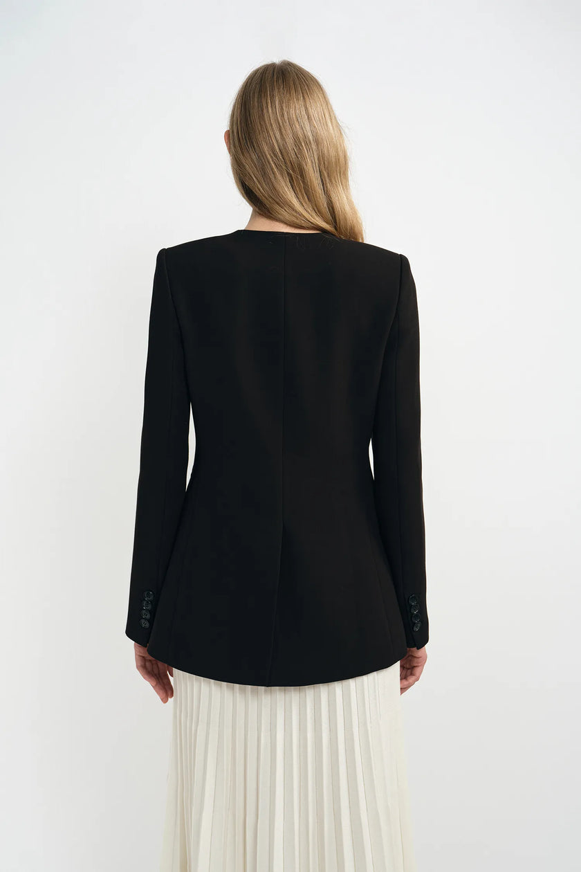 A model wearing a black collarless jacket, back view, showing a clean tailored silhouette.