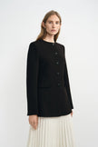 A model wearing a black collarless jacket with structured shoulders, three-quarter side view.