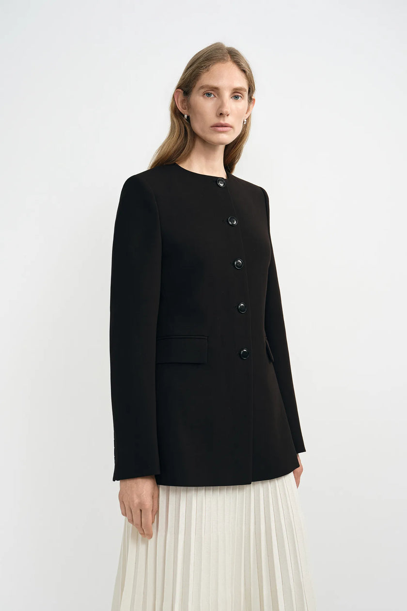 A model wearing a black collarless jacket with structured shoulders, three-quarter side view.