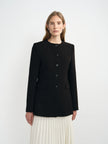 A woman modeling a black long-sleeve jacket with a button front, paired with a cream pleated skirt, against a plain light gray studio background.