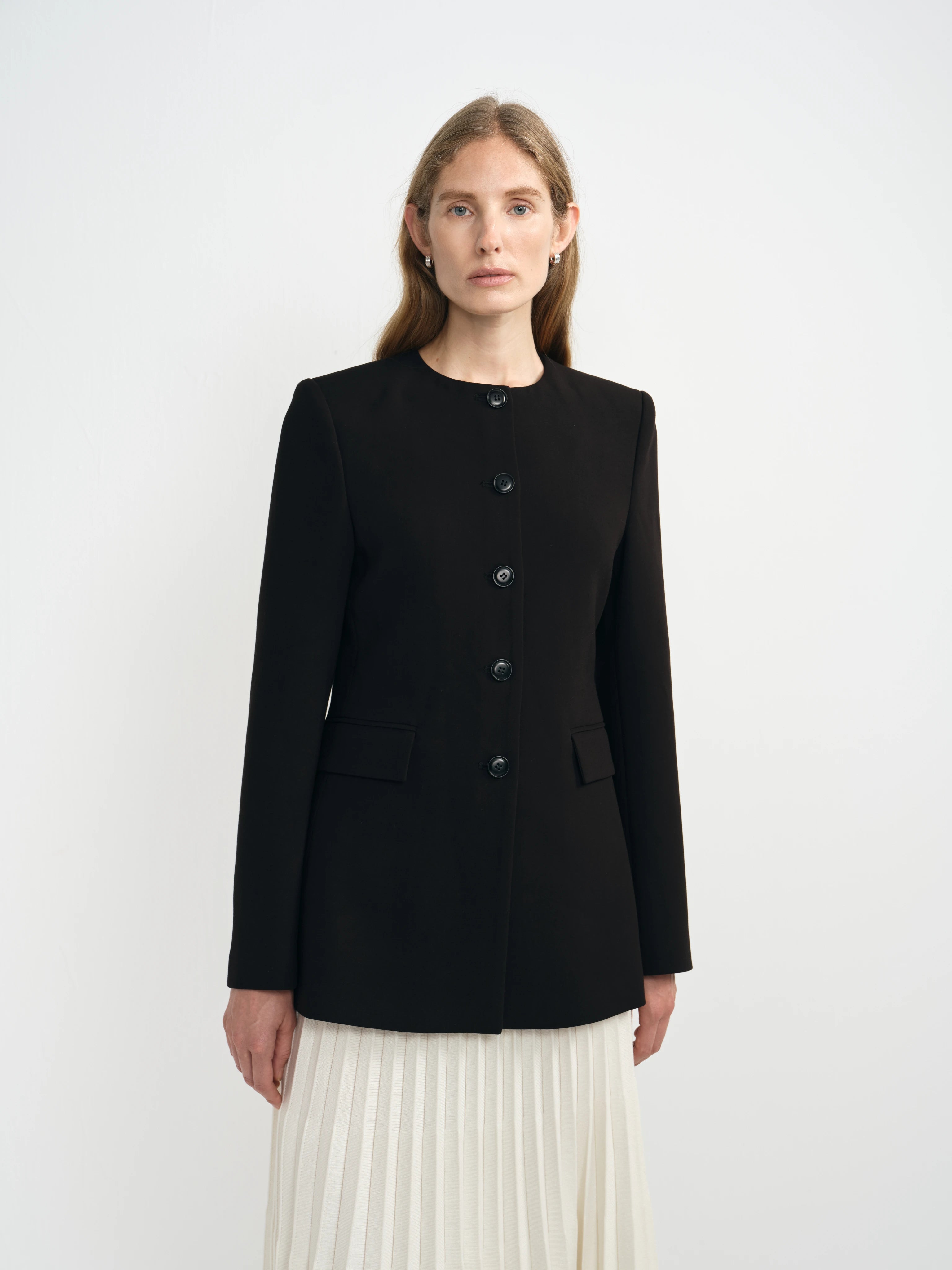 A woman modeling a black long-sleeve jacket with a button front, paired with a cream pleated skirt, against a plain light gray studio background.