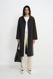 A model stands against a light gray studio background wearing a long dark brown coat over a white top and cream wide-leg trousers, holding a black tote bag.