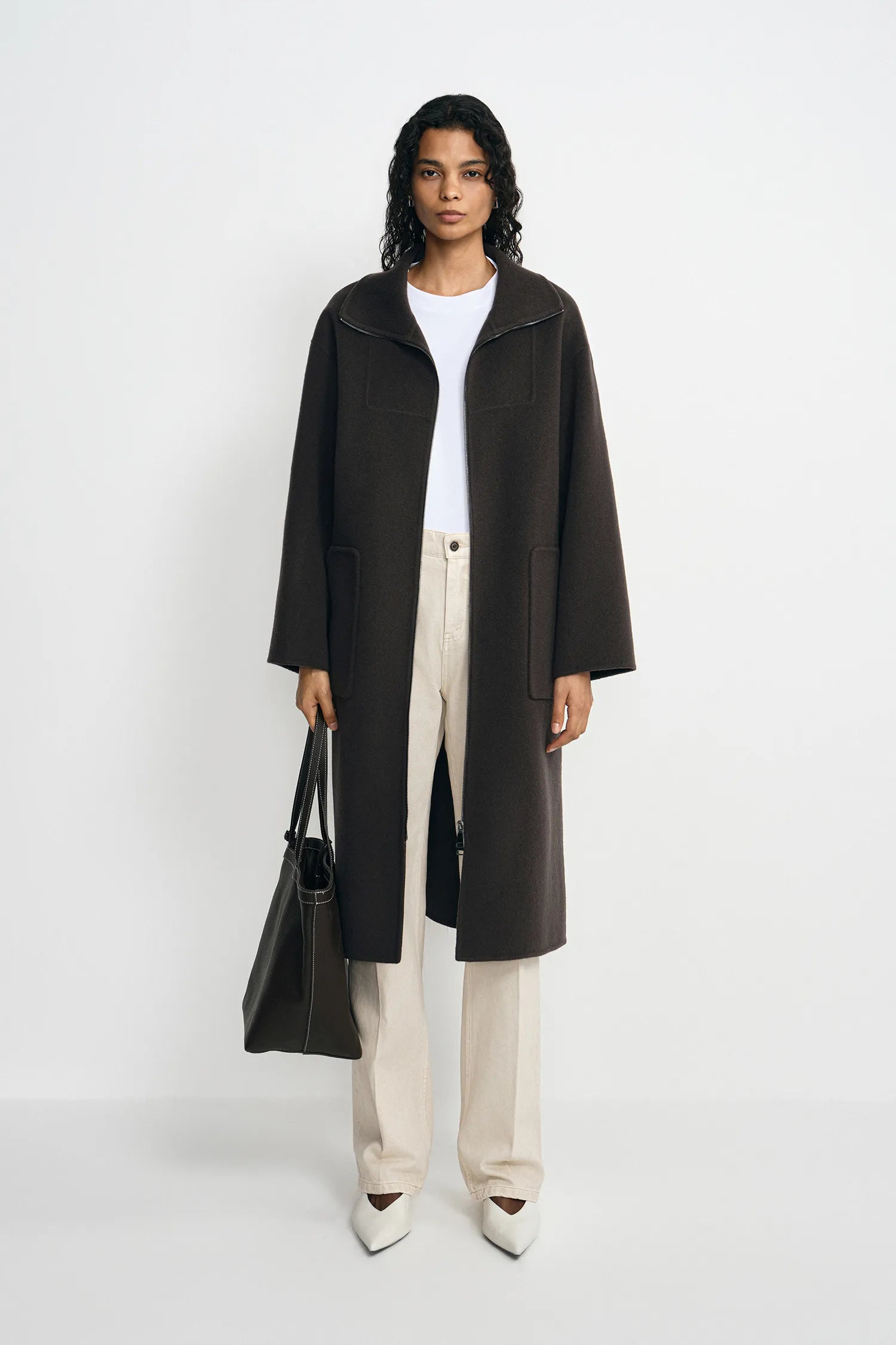 A model stands against a light gray studio background wearing a long dark brown coat over a white top and cream wide-leg trousers, holding a black tote bag.