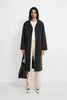 A model stands against a light gray studio background wearing a long dark brown coat over a white top and cream wide-leg trousers, holding a black tote bag.