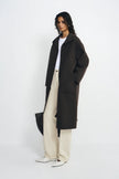 A model stands in a studio wearing a long dark brown coat over a white top and cream wide-leg trousers, holding a black bag against a plain light gray background.