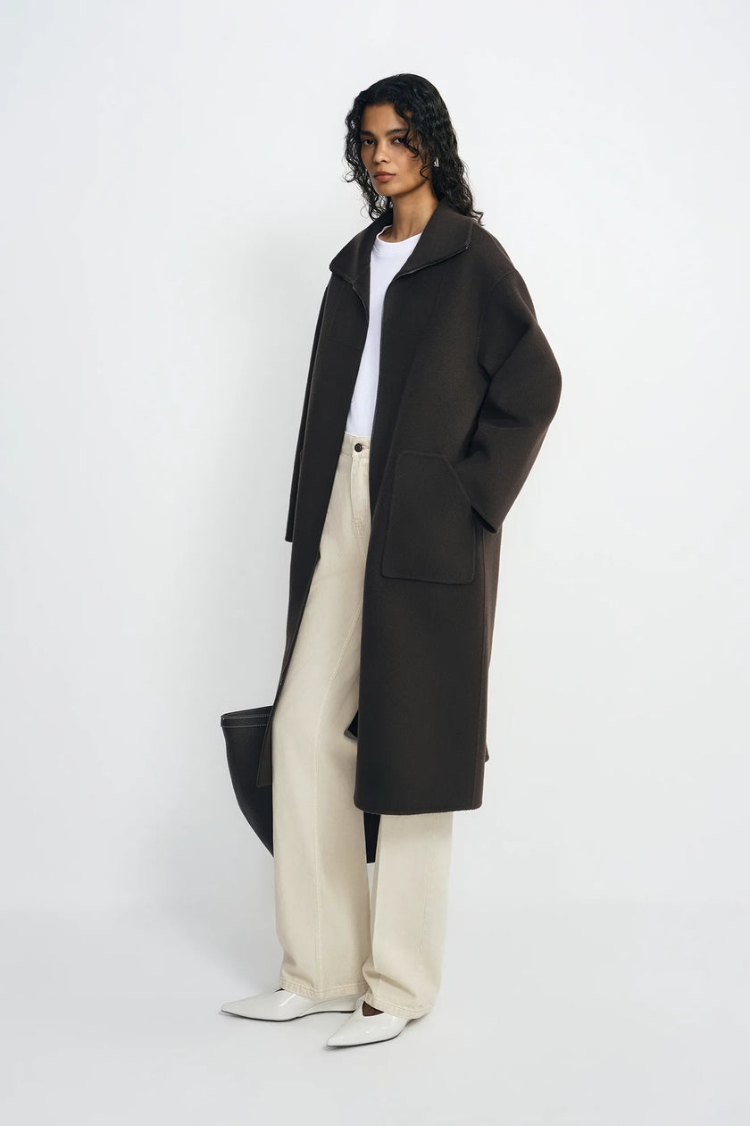 A model stands in a studio wearing a long dark brown coat over a white top and cream wide-leg trousers, holding a black bag against a plain light gray background.
