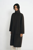 A model wearing a dark brown, mid-length coat with a high mock neck and large front patch pockets, paired with off-white trousers, against a plain light gray background.