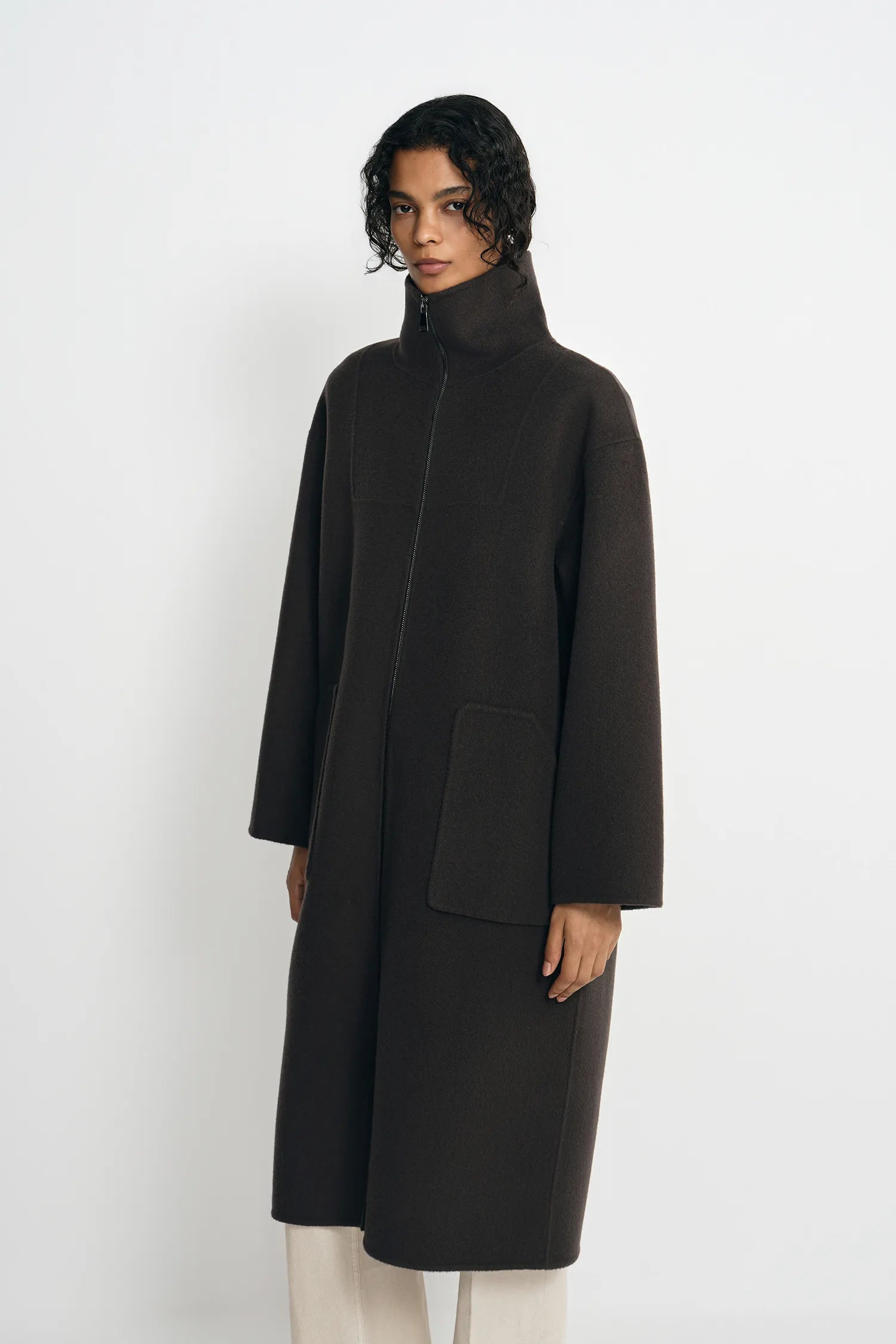 A model wearing a dark brown, mid-length coat with a high mock neck and large front patch pockets, paired with off-white trousers, against a plain light gray background.