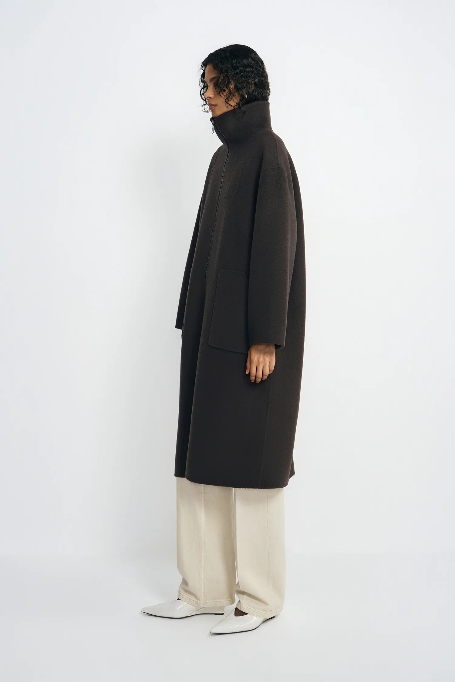 A model is shown in profile wearing a long, dark brown turtleneck coat, cream wide-leg trousers, and white pointed shoes against a plain white studio background.