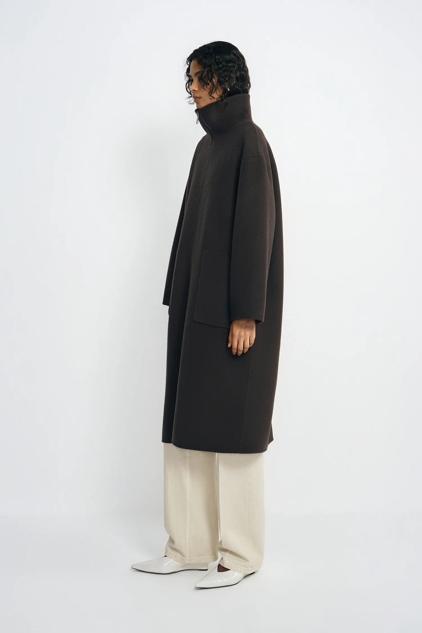 A model is shown in profile wearing a long, dark brown turtleneck coat, cream wide-leg trousers, and white pointed shoes against a plain white studio background.