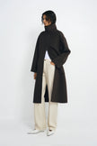 A model wearing a long, oversized dark brown turtleneck coat over a white top and cream wide-leg trousers, standing against a plain white studio background.