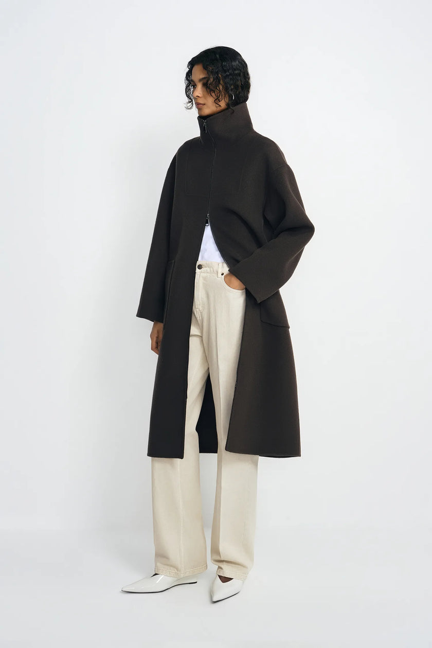 A model wearing a long, oversized dark brown turtleneck coat over a white top and cream wide-leg trousers, standing against a plain white studio background.