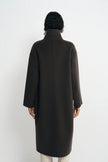 A person shown from the back wearing a long dark brown coat with a high mock neck, photographed against a plain light gray studio background.