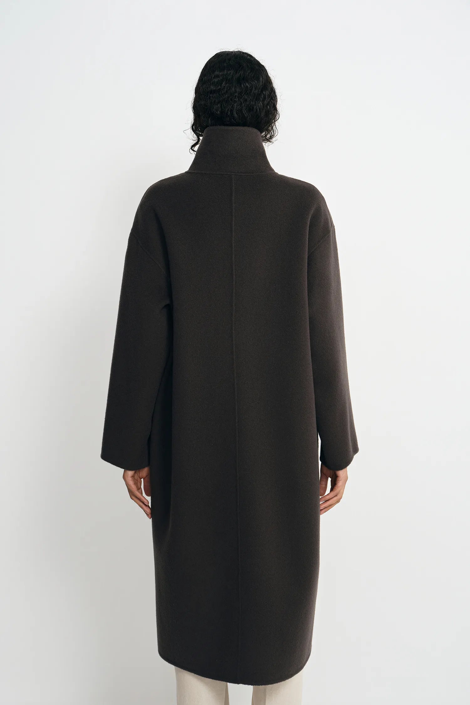 A person shown from the back wearing a long dark brown coat with a high mock neck, photographed against a plain light gray studio background.
