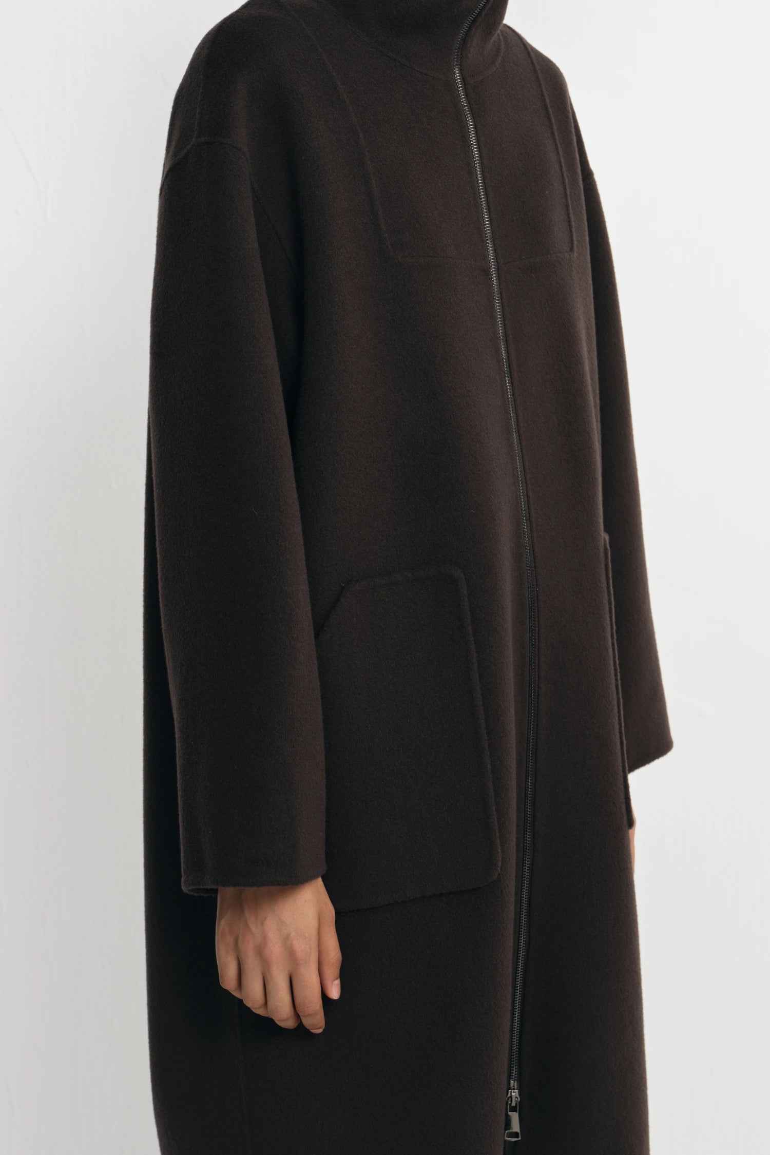 A dark brown wool-blend coat with a high mock neck, front zipper, and large patch pockets, shown on a model against a light gray background.