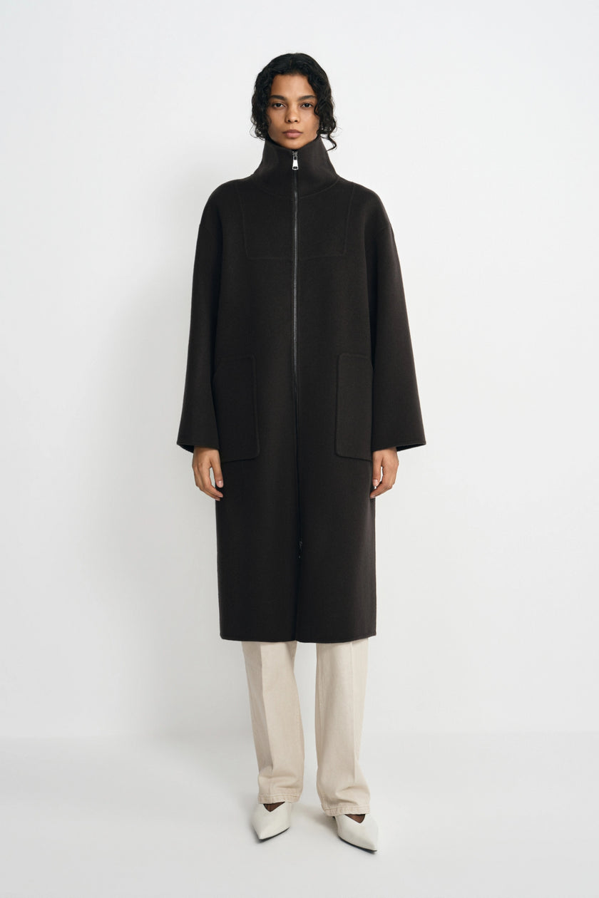 A model stands against a light gray studio background wearing a long black coat with a high mock neck and front zipper, paired with light beige trousers and white pointed shoes.