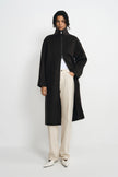 A model stands against a light gray studio background wearing a long black coat with a high mock neck and front zipper, styled over a white top and cream wide-leg trousers.