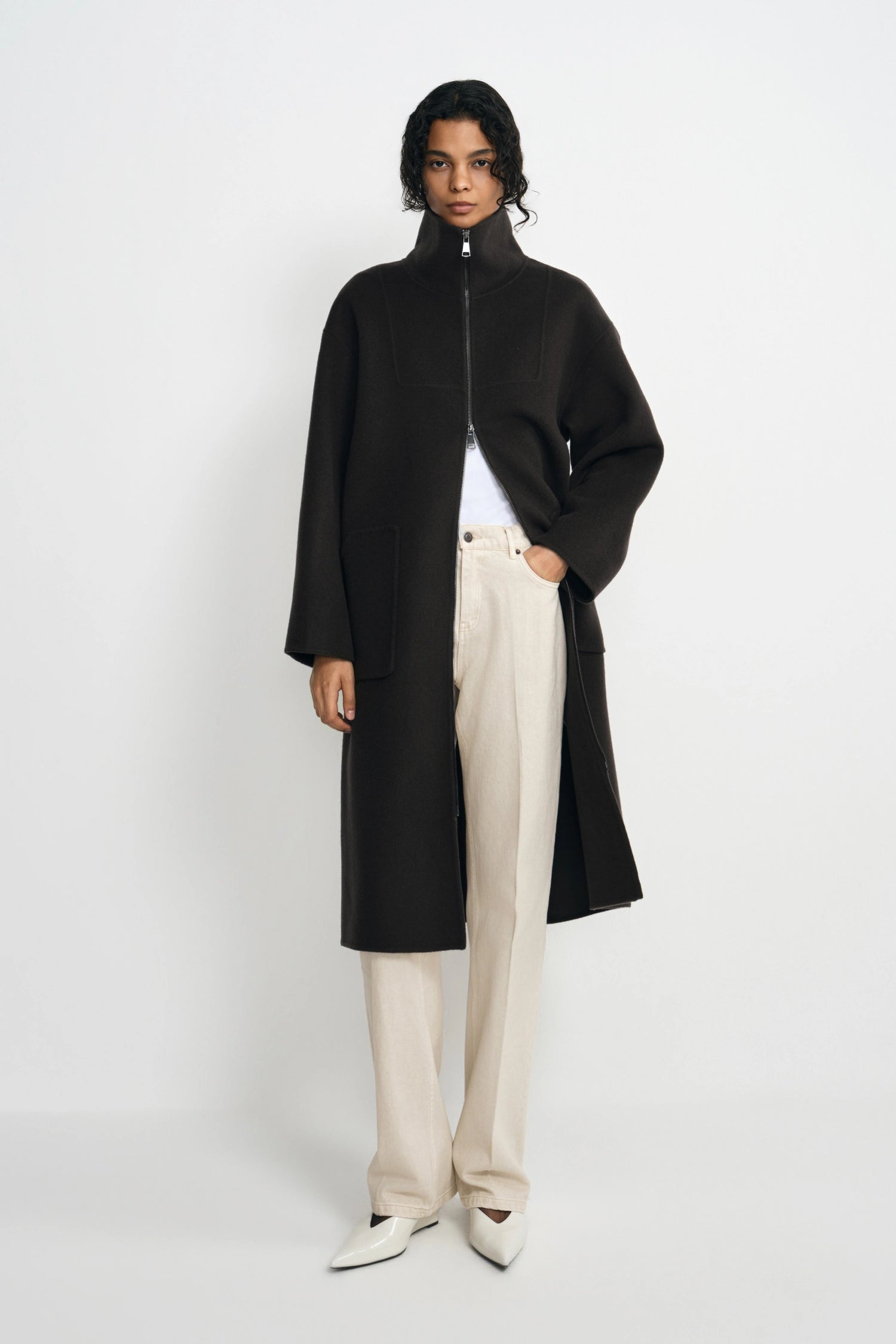 A model stands against a light gray studio background wearing a long black coat with a high mock neck and front zipper, styled over a white top and cream wide-leg trousers.