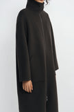 A person wearing a long black turtleneck coat with a front zipper and large patch pockets, shown in a side profile against a light gray studio background.