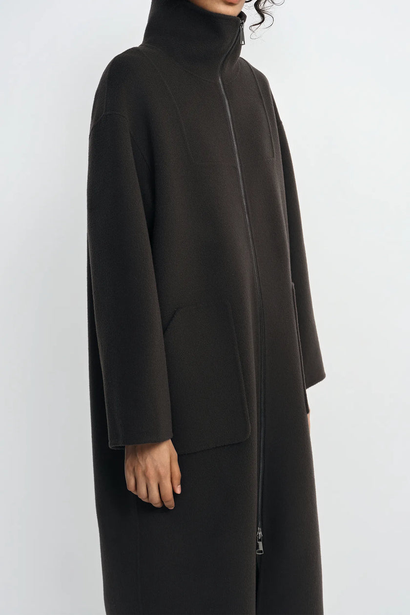 A person wearing a long black turtleneck coat with a front zipper and large patch pockets, shown in a side profile against a light gray studio background.