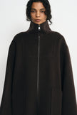 A model wearing a dark brown high-neck zip-up coat with a high collar and front pockets, photographed against a light gray studio background.