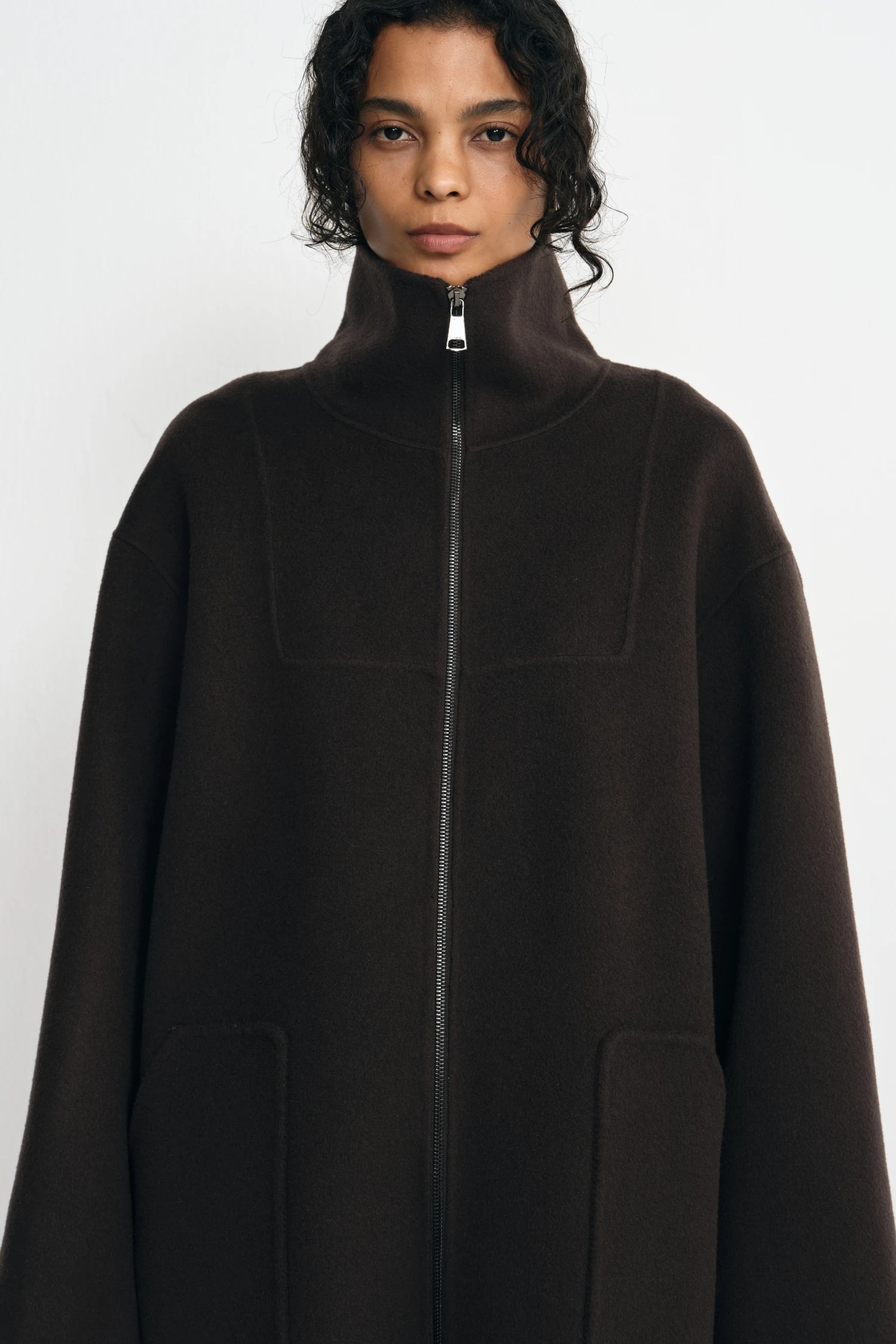 A model wearing a dark brown high-neck zip-up coat with a high collar and front pockets, photographed against a light gray studio background.