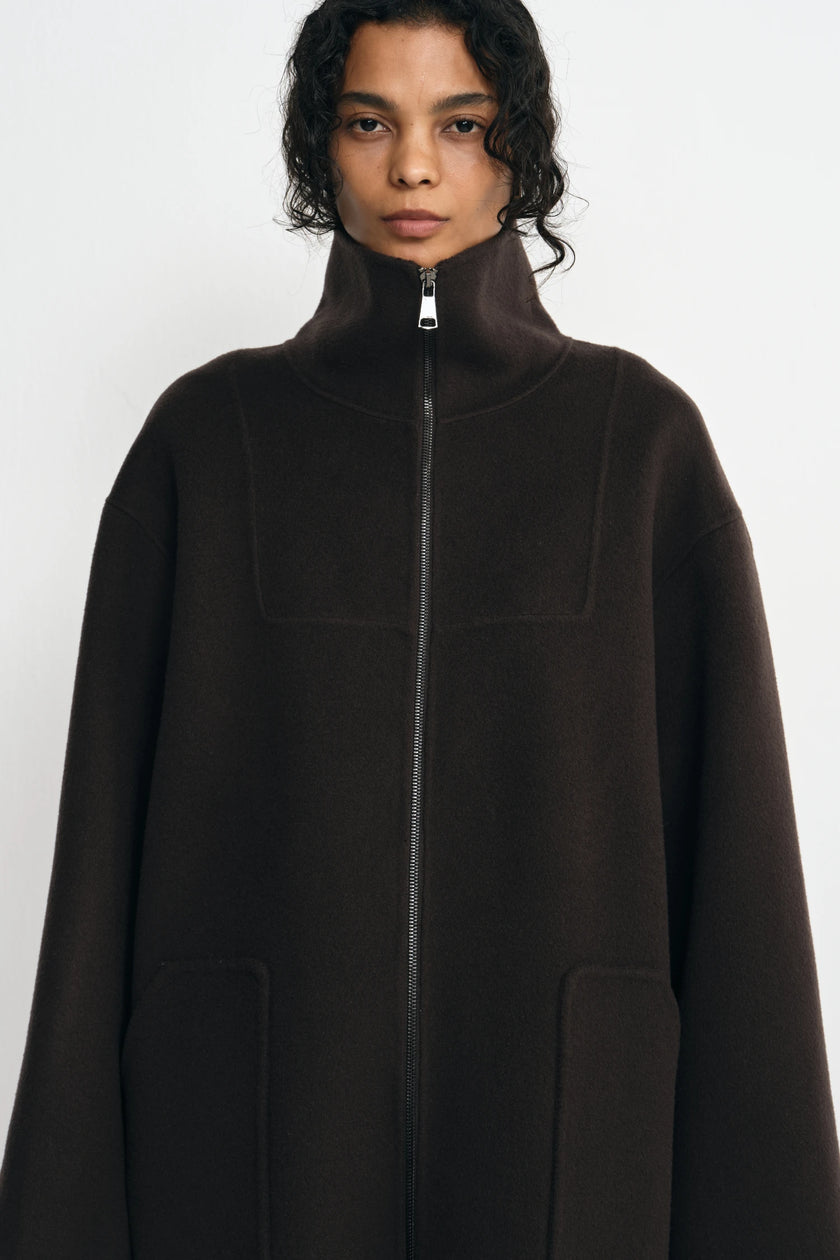 A model wearing a dark brown high-neck zip-up coat with a high collar and front pockets, photographed against a light gray studio background.