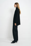 A woman stands in profile wearing an oversized black turtleneck blazer and matching wide-leg black trousers with black pointed heels, photographed against a clean white studio background.
