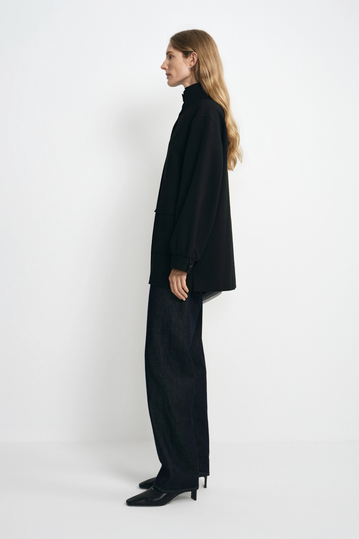 A woman stands in profile wearing an oversized black turtleneck blazer and matching wide-leg black trousers with black pointed heels, photographed against a clean white studio background.