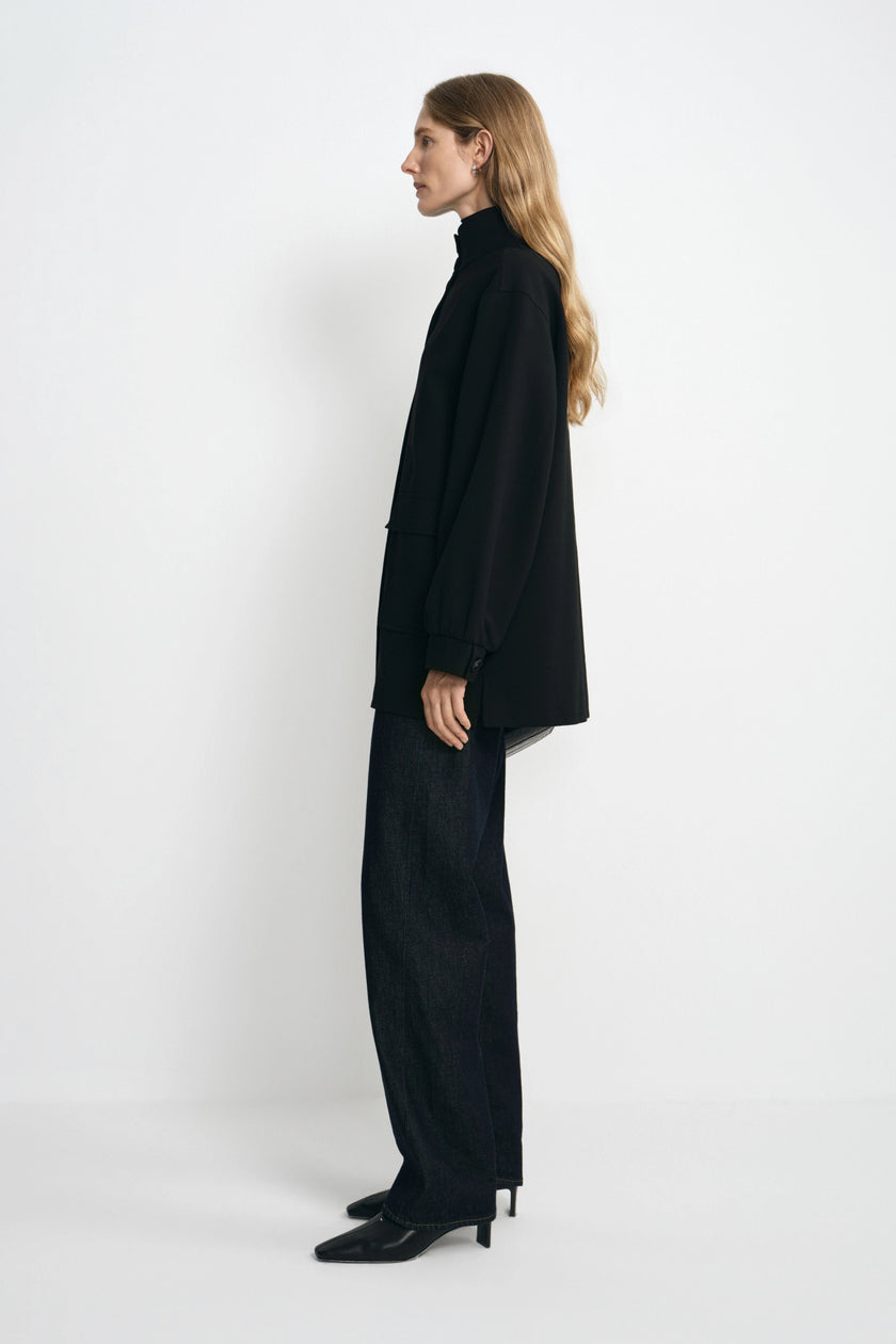 A woman stands in profile wearing an oversized black turtleneck blazer and matching wide-leg black trousers with black pointed heels, photographed against a clean white studio background.