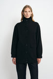 Front view of a person wearing an oversized black high-neck button-front jacket with flap patch pockets, layered over a black turtleneck and dark trousers, shown against a plain light background
