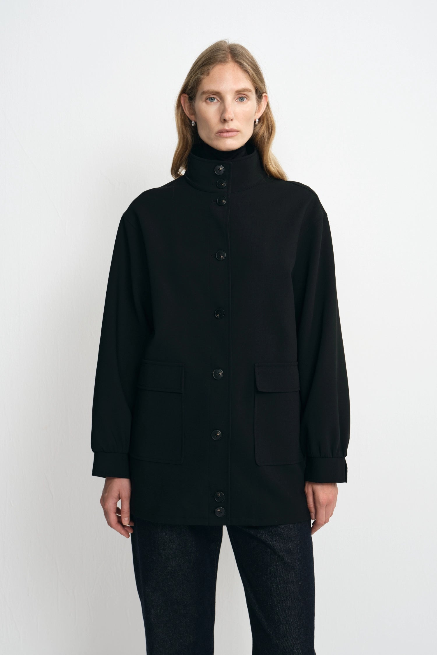 Front view of a person wearing an oversized black high-neck button-front jacket with flap patch pockets, layered over a black turtleneck and dark trousers, shown against a plain light background
