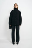 A model stands against a light gray studio background wearing a black high-neck button-front jacket with flap pockets, black wide-leg trousers, and black pointed shoes, while holding a small black handbag.