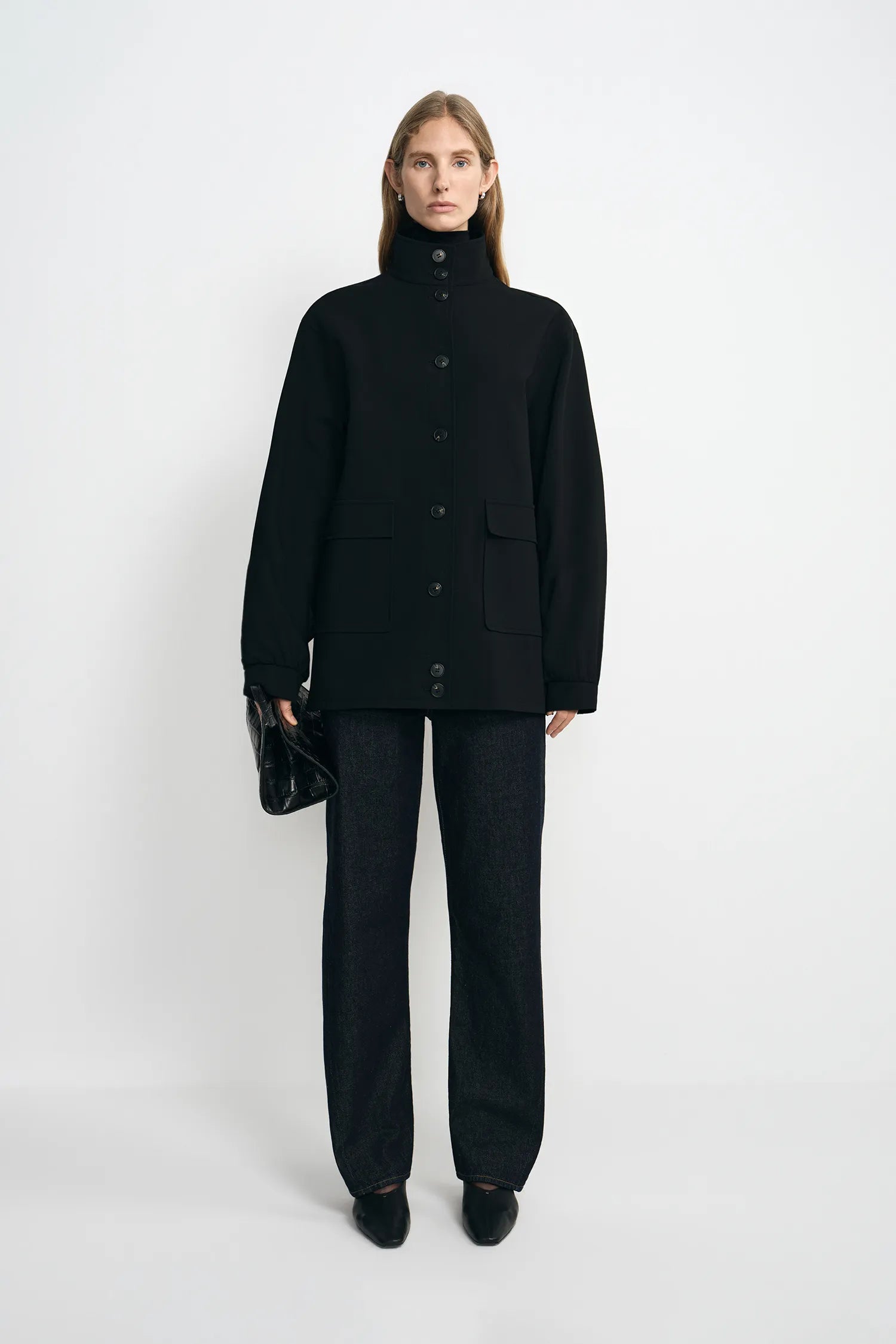 A model stands against a light gray studio background wearing a black high-neck button-front jacket with flap pockets, black wide-leg trousers, and black pointed shoes, while holding a small black handbag.